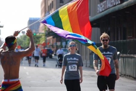 A woman and man in matching running shirts walk down a street as a rainbow flag flags in the foreground.