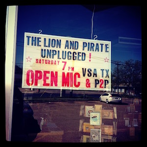 Photographed through a floor-to-ceiling, a marquee reads The Lion and Pirate Unplugged! Saturday 7pm OPEN MIC VSA TX & P2P. The lettering is black and red.