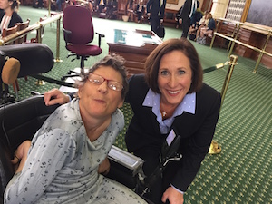 Two women pose for a photo, one sitting in a power chair with her arm tucked behind her, and the other leaning down to be shoulder-to-shoulder with the first.