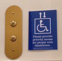 Next to the gold colored up and down buttons of an elevator, a blue sign with a handiman image reads Please provide priority access for people with disabilities.