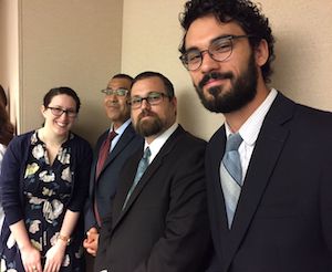 Four people in business dress are lined up casually against a wall, all turning to smile at the camera. 