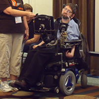 A man sits in a power chair, eyes cast down at a screen, while a microphone is held up to his speaking device.