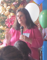 In a room with balloons and a decorated Christmas tree, a woman addressing a seated audience holds a microphone to speak while gesturing with her hand.