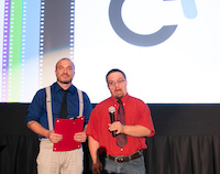 Garret, in a bright red shirt and tie, appears to be making a serious statement into the microphone he's holding. Beside him, Orion holds a folder with both hands and looks out into the audience with a wistful expression.