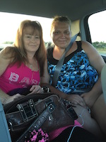 An older and younger woman sit side by side in the back seat of a car. They lean in towards each other and smile for the camera.
