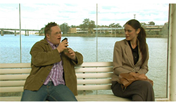 A man speaking into a tape recorder lounges on a bench, while the woman sitting next to him seems intrigued but hesitant.