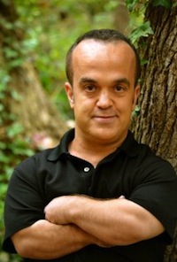 In a wooded area, a man with folded arms looks at the camera with an understated smile.