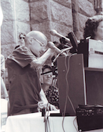 In a black and white photo shot from a side, a young man with muscular dystrophy looks down at a table with several microphones pointed at his face.