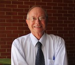With a red brick wall in the background, an older man in a button-up shirt and tie and glasses smiles.