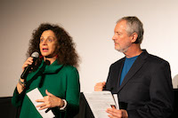 A woman with an adamant expression speaks into a microphone while a man next to her listens somberly.