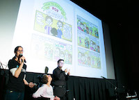 A comic book spread is projected onto the theater screen. A woman in the foreground speaks into a microphone, a woman seated next to her looks up at the screen, and a man next to then signs ASL.