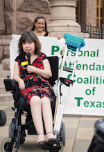 A woman in a summery red and black dress leans on the arm rest of her power chair and holds a microphone to her mouth. Behind her, a smiling woman holds up one side of a large banner that reads Personal Attendant Coalition of Texas.