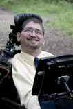A man in a power chair equipped with a screen grins at the camera.
