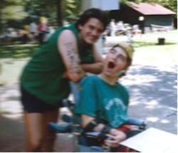 Two men, one sitting in a power chair, the other leaning on the back of the chair, smile at the camera