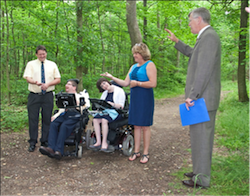 A group of 5 formally dressed people stand/ sit in the middle of a wooded area.