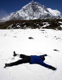 On snow-covered ground, with a mountain peak in the background, a warmly-dressed man with a prosthetic leg makes a snow angle. 