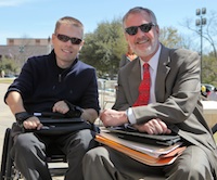 Two men in professional dress and sunglasses sit in a bright outdoor environment with tablets and stacks of folders in their laps. 