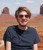 A man in sunglasses smiles at the camera with a sweeping desert landscape in the background