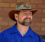 In front of a brown brick wall, a bearded man in a blue collared shirt and cowboy hat smiles in amusement at something off camera.