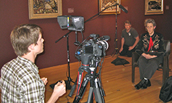 A woman in a western shirt sits in a chair and gazes off camera, while a young man in the fore-ground looks into the viewing screen of a video camera.