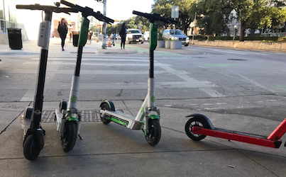 4 electric scooters stand in the middle of a curb cut, blocking the path to a crosswalk.