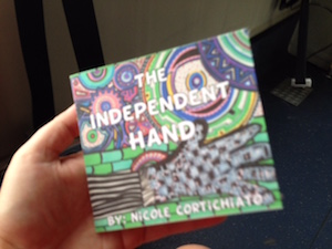 A hand holds up a book with a colorful cover in a hand drawn style to the camera. It reads The Independent Hand by Nicole Cortichiato.