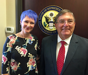 A woman with short periwinkle hair stands shoulder to shoulder with an older man in a suit. A US House of Representative seal is hung behind them.
