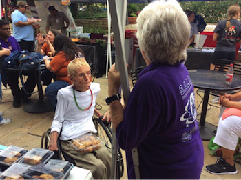 Amid groups of people sitting at outdoor tables and managing booths, a woman sits in a push chair with a clear box of cookies on her lap. In the foreground, a woman in a purple shirt with her back to the camera leans on a tent post.
