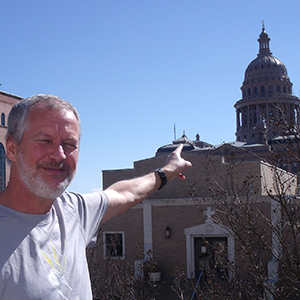 A man in a Lightning Laces running shirt smiles at the camera and points to the Texas Capital building in the background.