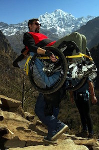 With a snow capped mountain in the back ground, two men ascend a rocky path. One is leaning back in a push wheelchair, strapped by a yellow cord to the back of the other.