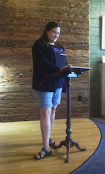 In a wood paneled corner resembling a stage, a woman reads off a podium.
