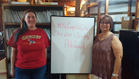 Two women stand on either side of a whiteboard that reads #NoCapsNoCuts to our clients' Medicaid. In the background is a large bookcase full of files.
