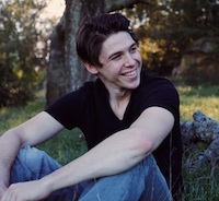 A man sits in an outdoor, wooded area, elbows on knees, grinning at something over his shoulder.