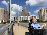 A man in a power chair rolls across a sidewalk with Austin's skyline and a bright blue sky behind him.