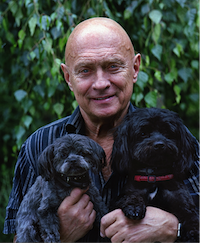 With green foliage in the background, an older man smiles directly at the camera and holds two small black dogs in his arms.