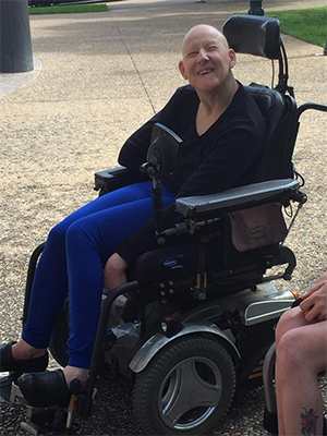 On a broad sidewalk outdoors, a bald woman in a power chair smiles happily and quints at the camera.