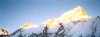 A wide photo of snow-capped mountain peaks, with bright sun shining off the very top.