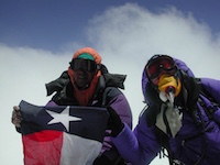 Against a bright blue sky with a wispy cloud, two people hold up a Texas flag. They are both heavily clothed with goggles obscuring their faces, and one also wears what appears to be an oxygen mask.