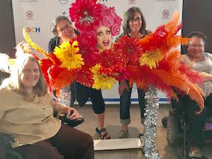 In front of a ZACH theater step and repeat banner, a space for photos has been set up with a cut-out of a pink-haired drag queen's smiling face surrounded by orange, red, and yellow feathers and flowers. Two women sitting in wheelchairs are in front of the display and two other women stand behind it, one sticking her tongue out.