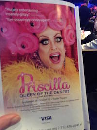A hand holds open a program booklet to a page where a made up drag queen with a pink curly headpiece and orange feather boa opens her mouth in a big smile toward the viewer. The words Priscilla Queen of the Desert appear at the bottom of the page. 