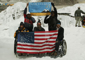 On a bright, snowy field, three warmly clothed men sitting in wheelchairs hold up a flag, which resembles the American with the field of stars replaced by a figure in a wheelchair. Behind them, two more bundled up people stand, holding up a smaller banner that reads Team Everest 03