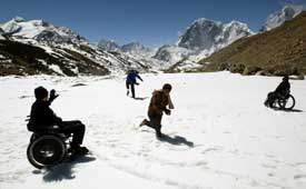 On a flat, snowy field with mountains in the background, four people play in the snow. One chases another on foot, while two in wheelchairs watch and wave from either side.