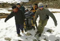 Over snow, four bundled up men carry a fifth in a wheelchair, each taking a corner of the chair.