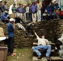Surrounded by onlookers, a man sits on the top step of a stone stairway. He hands a folded up wheelchair to a second seated man, reaching up and backward from the bottom step. 