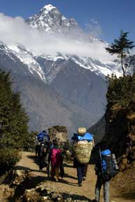 With a snow-capped mountain and clouds in the background, a large group of people with towering packs on their backs walk up a dirt trail.