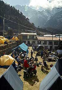 From a second-story perspective, a large group of people, several in wheelchairs, mill about among camping tents of various shapes and colors. A multi-story structure is behind them, and mountains and clouds loom further in the distance.