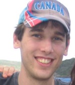 Close up on a grinning young man with ball cap that reads Canada. A hand is resting on his shoulder and mountains form the background.