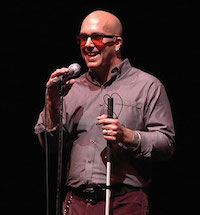 A smiling man with a white cane and red-lensed glasses holds a microphone up to his mouth.