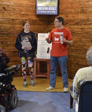 In the corner of a wood paneled room, a girl with cat leggings stands next to a man who is talking and gesturing with his hands. Both are holding sheets of paper.