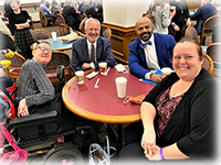 Four people sit around a cafeteria table, one in a power chair. All turn or lean in to smile at the camera.